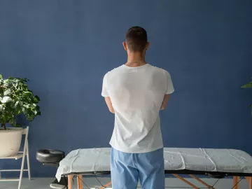 A massage therapist stands over a massage table, facing away from the camera in a pensive tone.