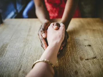 Two people grasping hands in a comforting manner across a table from each other.