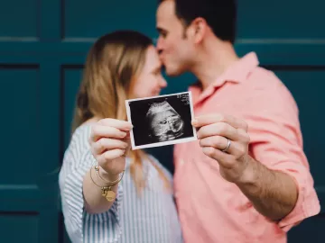 a male and female couple kiss while they hold up an ultrasound photo of a baby.