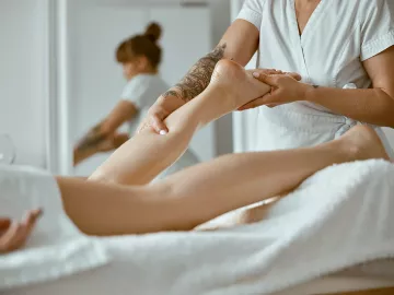 A massage therapist dressed in white works on a prone client's lower calf.