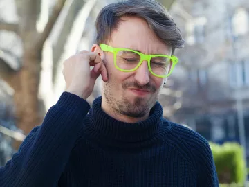 A man in a navy sewater and neon glasses clutches his ear and winces.