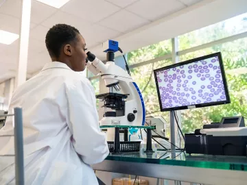 A young Black woman in a labcoat analyzing cells underneath a microscope.