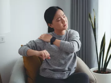 A woman sitting in a chair grabs her shoulder in pain.