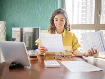 A woman smiles while looking at paperwork. A calculator and tablet are on the table in front of her.