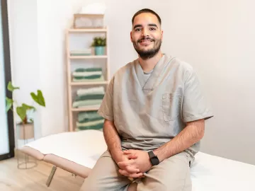A massage therapist sits on the edge of a massage table, smiling for a photo.