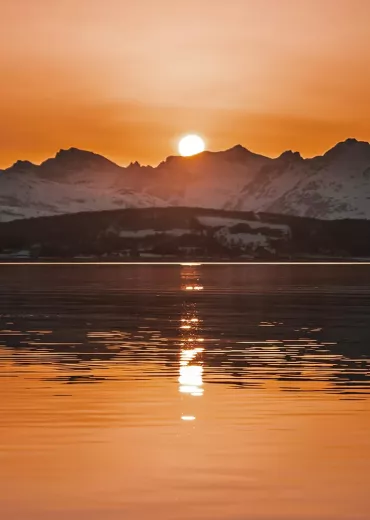 The sun sets over snowy peaks and a lake.