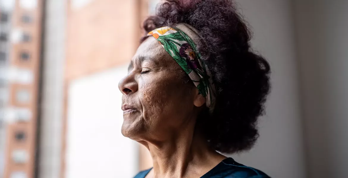 mature woman standing near a window with eyes closed breathing in.
